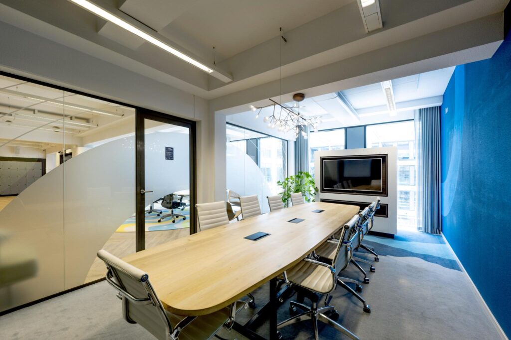 Modern conference room at Mr. Treublaan with a large wooden table, office chairs, and a wall-mounted screen.